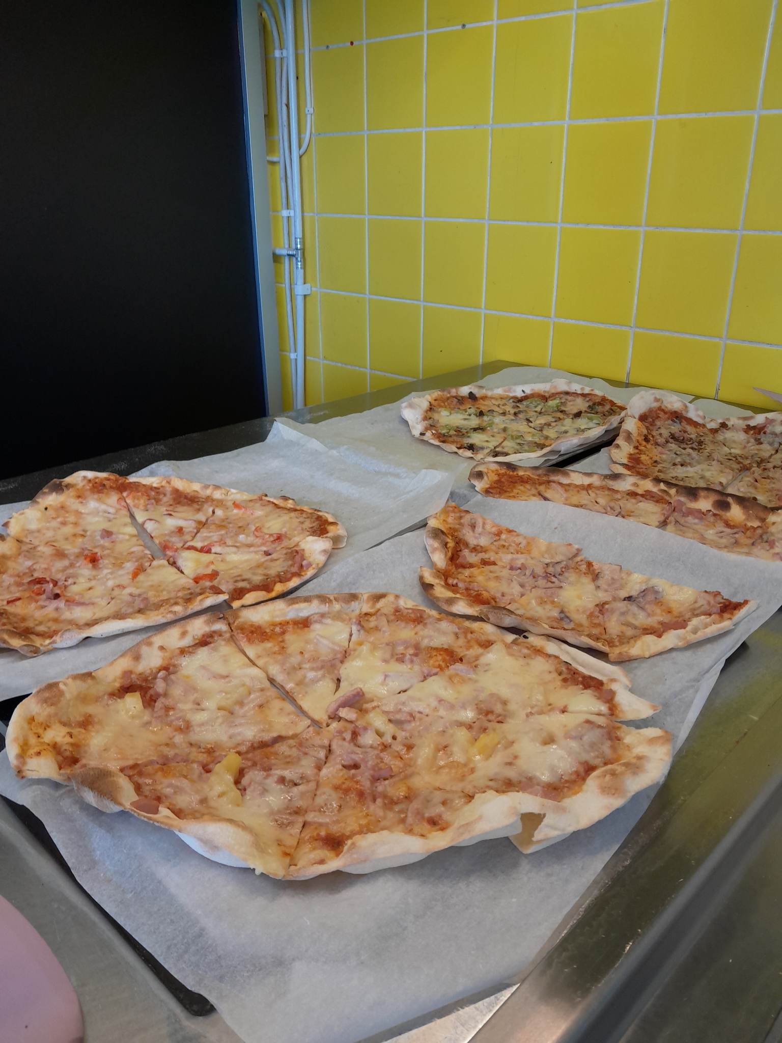 Several thin-crust pizzas and slices on parchment paper laid out on a stainless counter in front of a bright yellow tiled wall; cheese-covered with visible ham and pineapple-style toppings on some pies.
