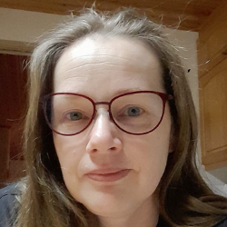 Close-up portrait of an adult woman with long brown hair and red-rimmed round glasses, neutral expression, soft lighting, standing in a home kitchen with wooden cabinets visible behind her.