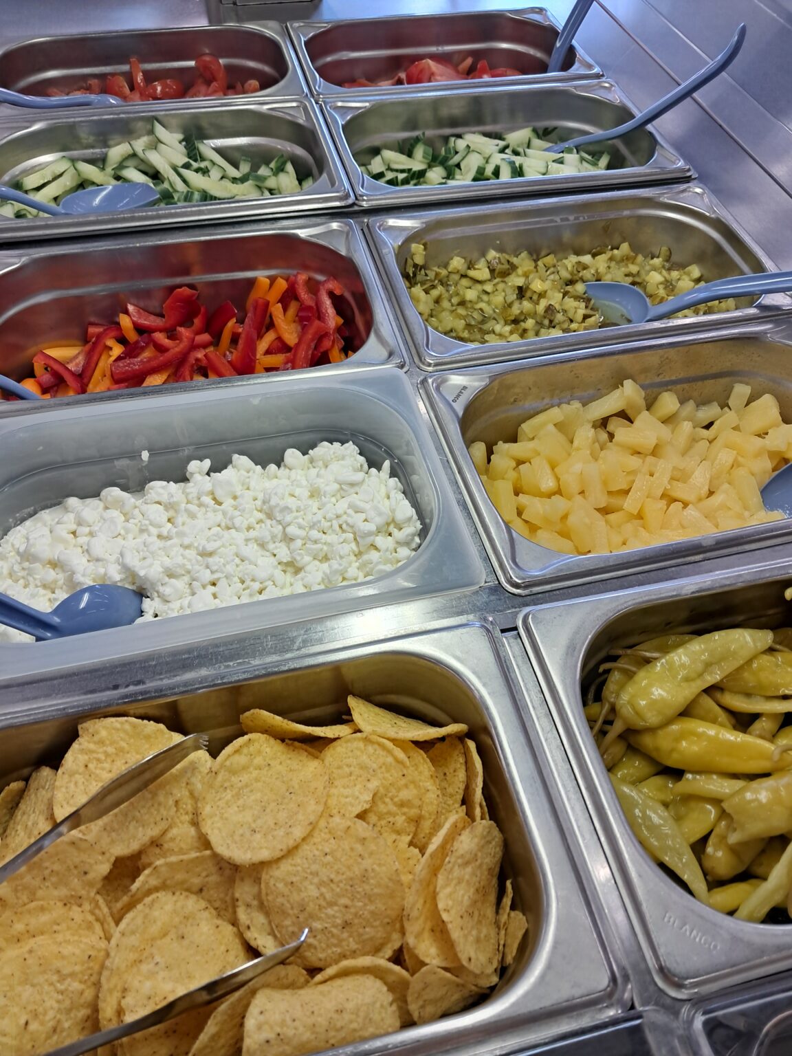 Stainless-steel salad bar with trays of chopped tomatoes, sliced cucumbers, red and yellow bell peppers, diced pickles, cottage cheese, pineapple chunks, tortilla chips and pickled peppers with serving spoons.