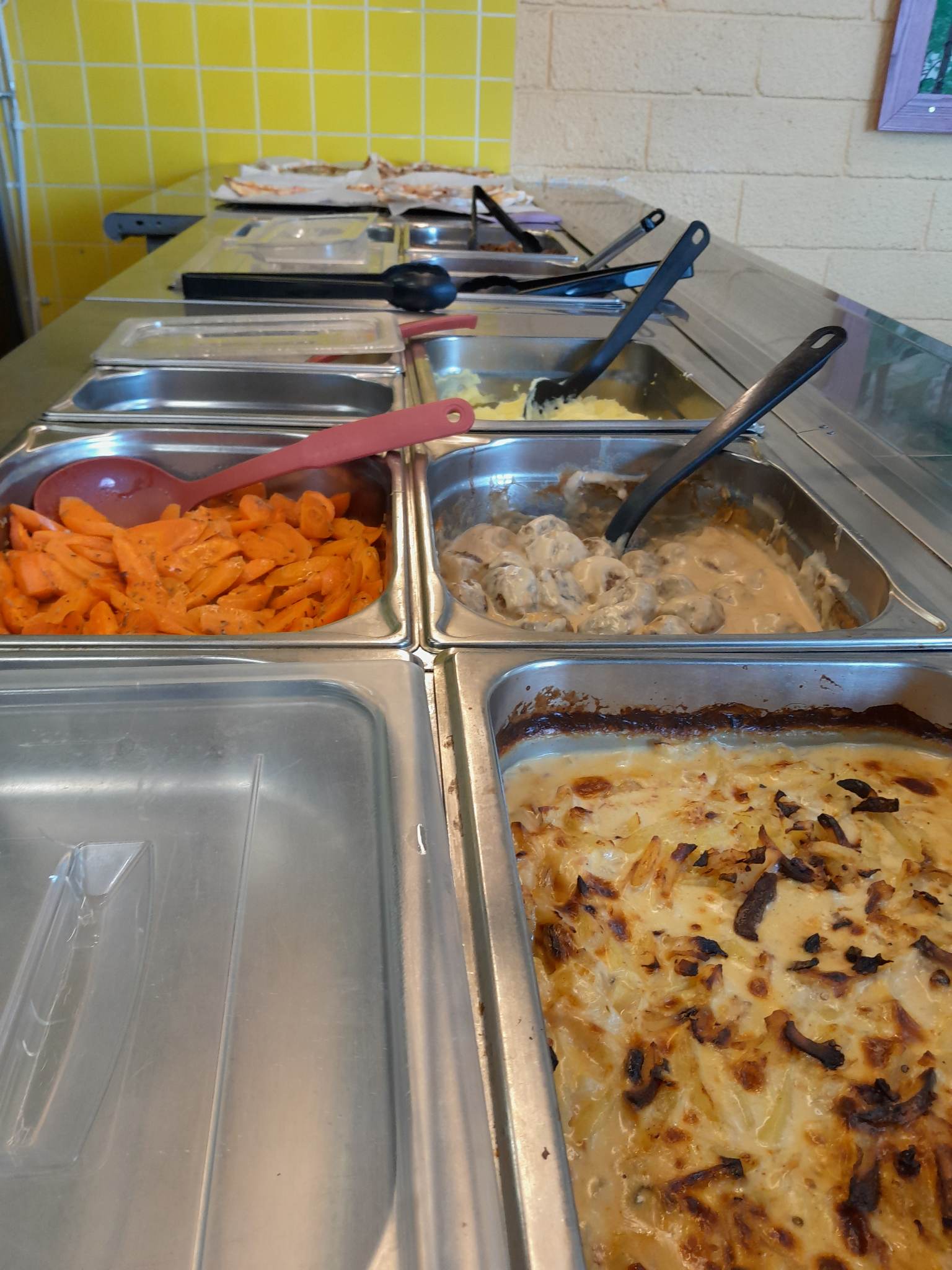 Stainless-steel cafeteria buffet with hot pans: glazed sliced carrots with a red spoon, meatballs in creamy gravy, mashed potatoes and a browned potato gratin, black utensils and a yellow tiled wall behind.