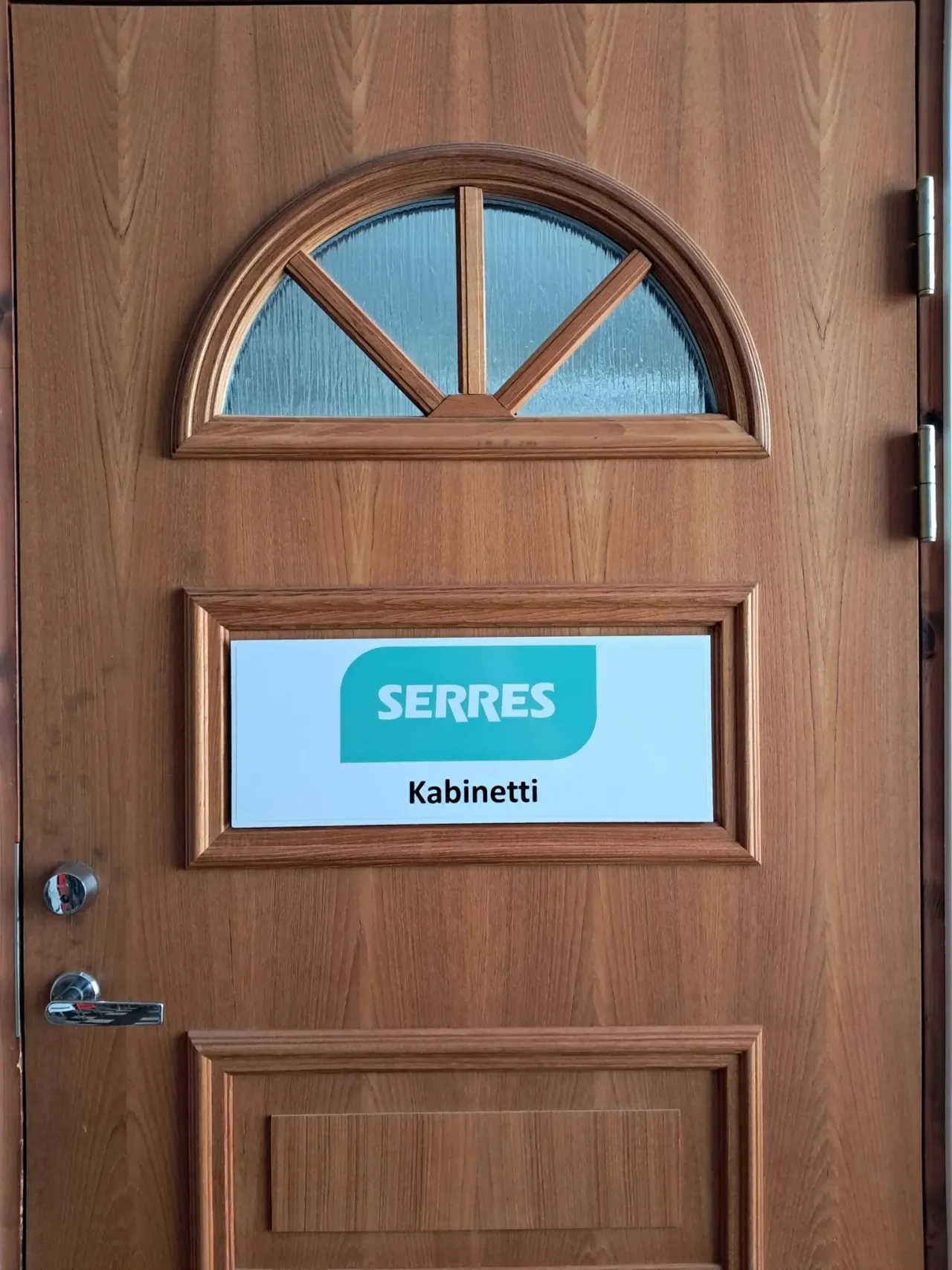 Wooden door with a semicircular divided glass window, framed recessed panels, chrome handle and hinges, and a white sign reading 'SERRES' with 'Kabinetti' below mounted in the middle panel.
