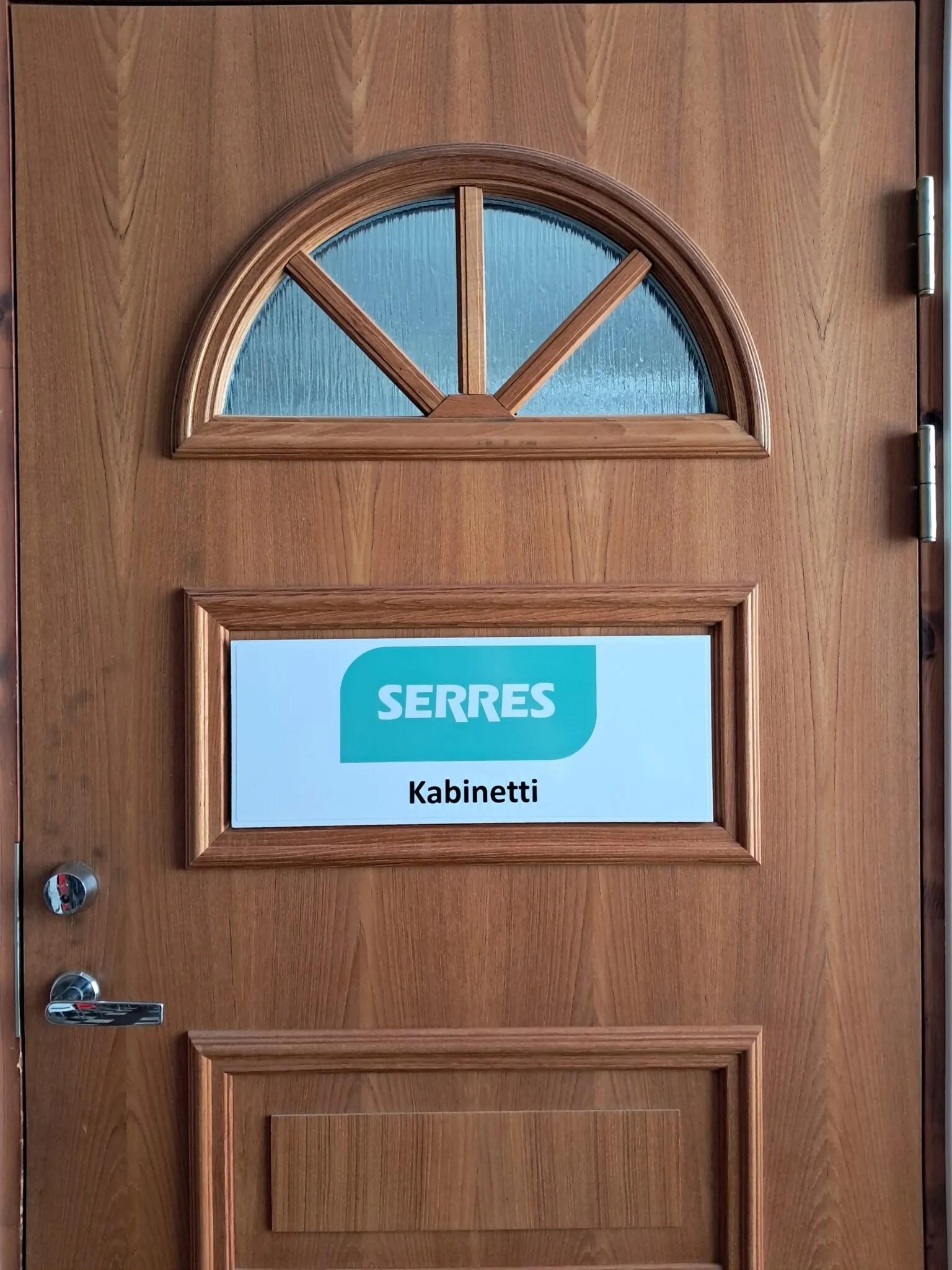 Wooden door with a semicircular divided glass window, framed recessed panels, chrome handle and hinges, and a white sign reading 'SERRES' with 'Kabinetti' below mounted in the middle panel.