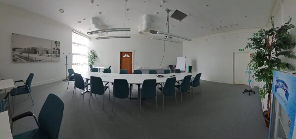 Empty modern conference room with U-shaped white tables and teal chairs, high white ceiling with exposed ducts and suspended lights, large window with vertical blinds, potted plants, flip chart and wooden door.