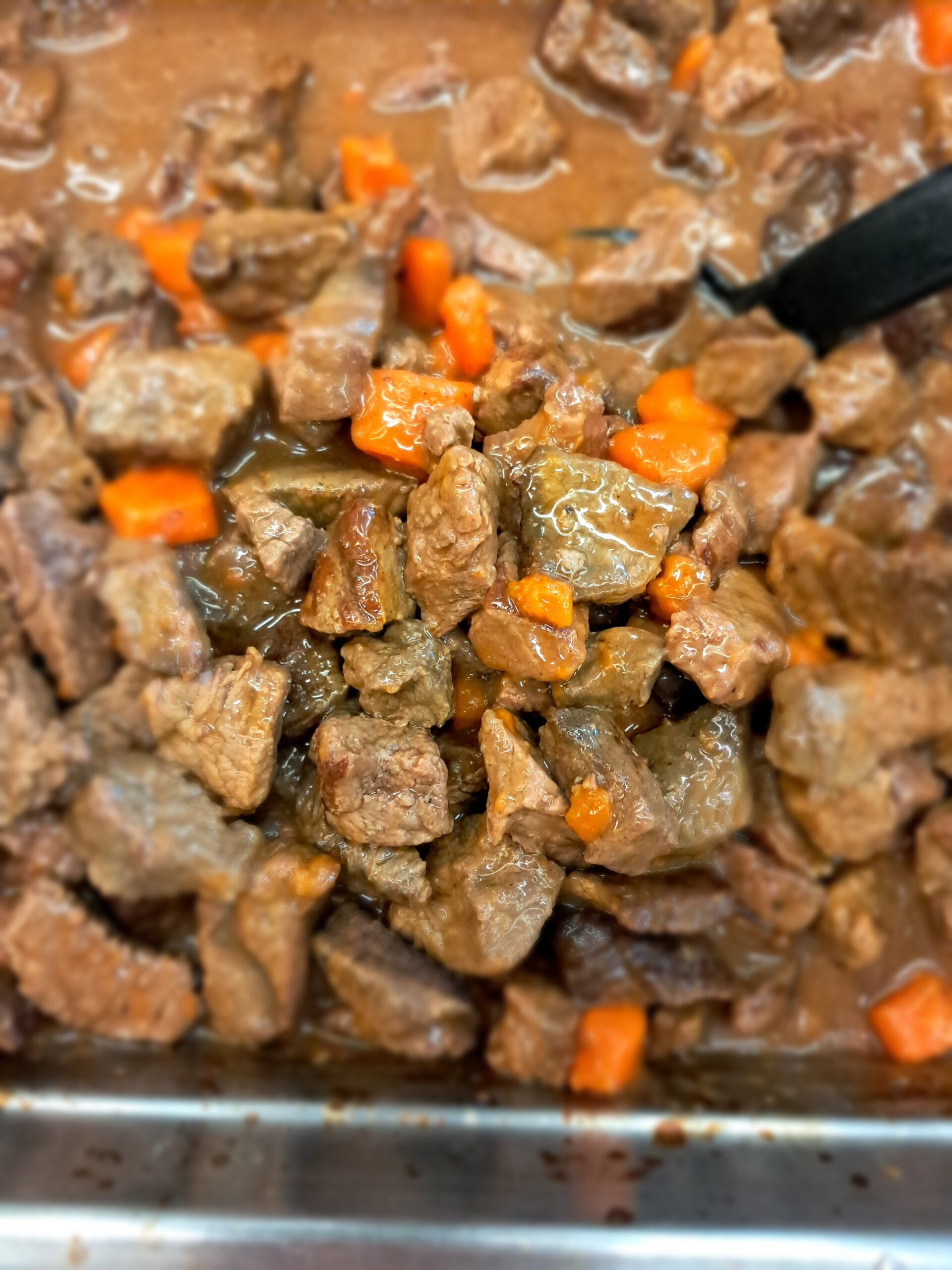 Close-up overhead view of a hearty beef stew: tender browned beef cubes and carrot chunks coated in rich brown gravy inside a stainless-steel serving tray.
